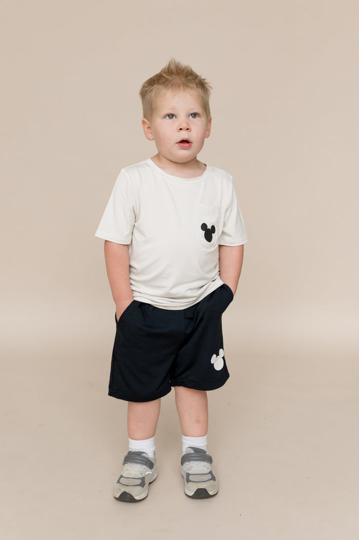 Young child wearing a white t-shirt and black shorts with a small mickey design on a beige background