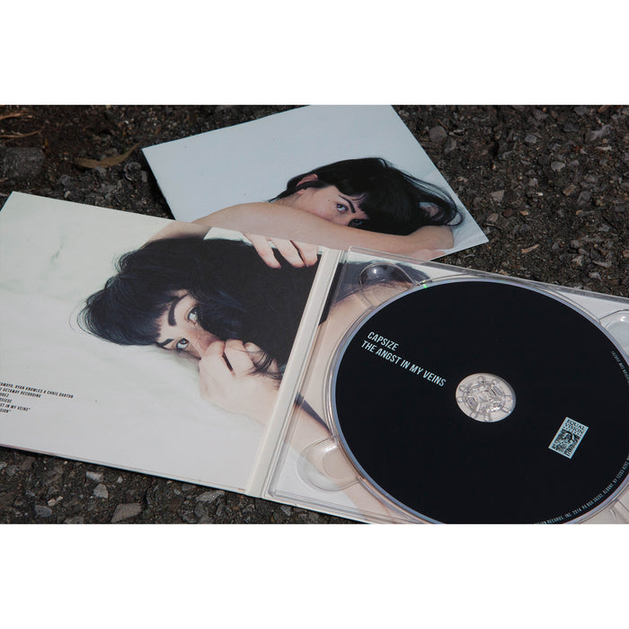 A CD case lies open on the ground, revealing a Capsize The Angst In My Veins CD and booklet featuring a woman with dark hair on a white surface, partially covering her face.