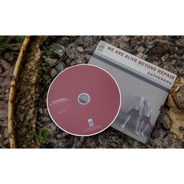 A maroon Gatherers CD sits on a rusty surface next to its cover, showing two people with their heads down beneath the title We Are Alive Beyond Repair, capturing the somber mood of Infinity & Gloom.