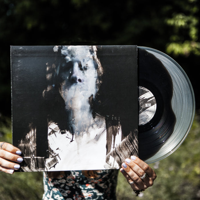 Someone holds a Capsize Limited Pressing vinyl and its cover in front of their face outdoors, perfectly aligning the abstract portrait on the sleeve with their head for a seamless blend of body and artwork against a green backdrop.