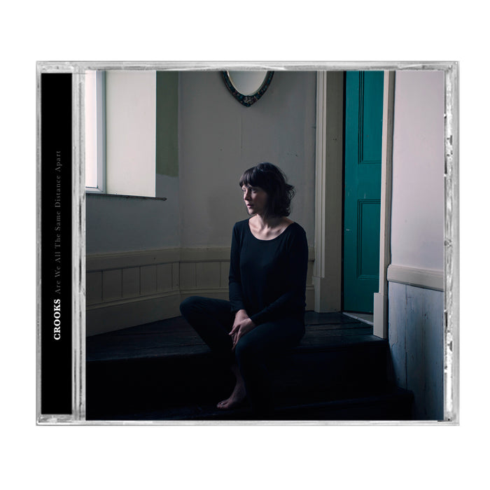 A Crooks UK CD case showcasing an album cover of a woman in black on dark wooden stairs, barefoot, in a minimalist room with white walls and a green door. Track list included inside.