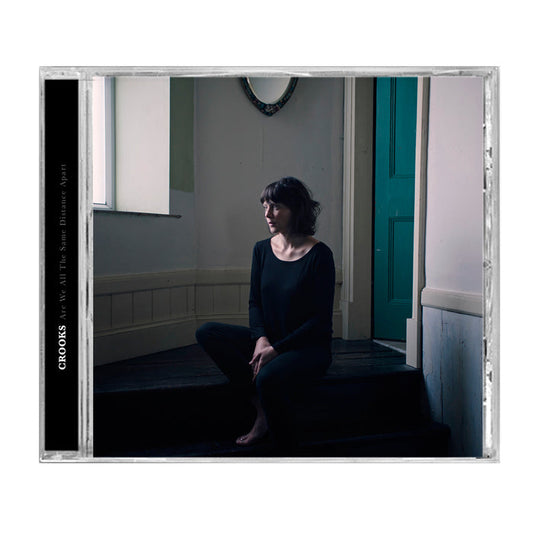 A Crooks UK CD case showcasing an album cover of a woman in black on dark wooden stairs, barefoot, in a minimalist room with white walls and a green door. Track list included inside.