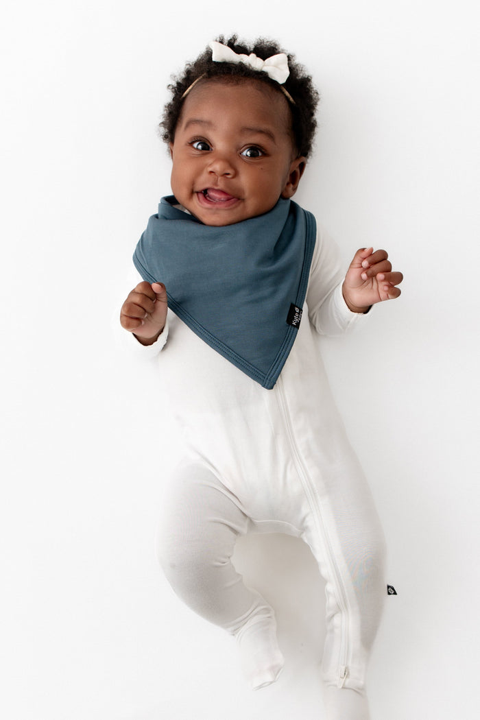 Infant girl laying down wearing the Bib in Atlantic paired with a Cloud Zippered Footie and Cloud Tiny Nylon Bow
