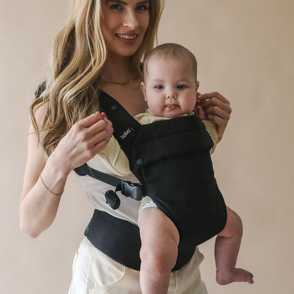 Woman holding a baby in a Boba Auri black baby carrier against a beige background
