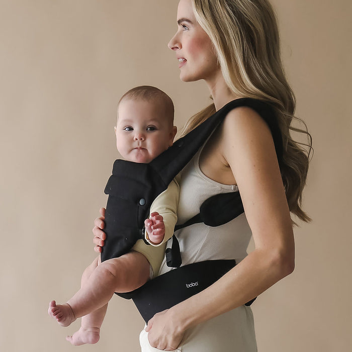 Woman holding a baby in a Boba Auri black baby carrier against a beige background