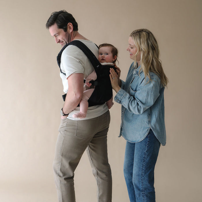 Man using a Boba Auri baby carrier and holding a baby, with a woman standing beside them against a beige background.