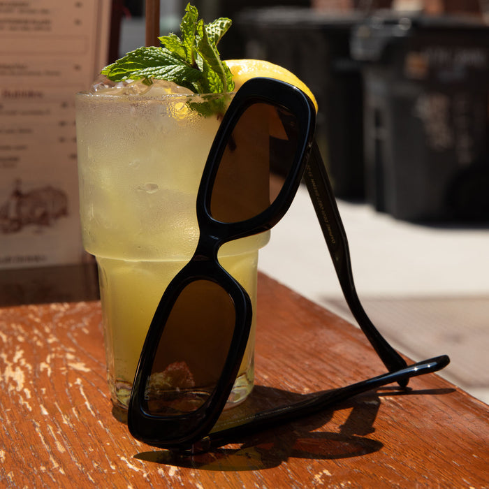 flay lay of diff eyewear featuring the daphne rectangle sunglasses with a black with silver metal frame and brown lenses leaning on a glass of lemonade.
