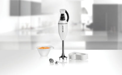 Der ESGE-Zauberstab 902105 Set E120 Select Stabmixer von ESGE Zauberstab steht aufrecht auf einer glänzenden Oberfläche neben einer Schüssel mit geschnittenem orangefarbenem Gemüse und verschiedenen Accessoires, im Hintergrund ist eine verschwommene moderne Küche zu sehen.
