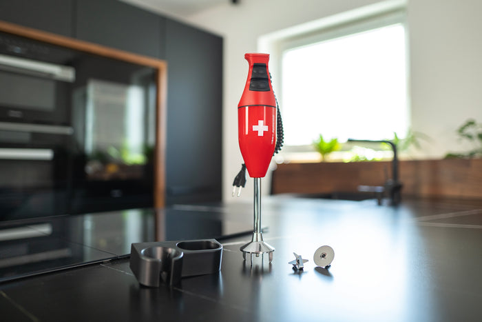 Das ESGE-Zauberstab E120 Select Premium-Set 902125, ein roter Stabmixer von ESGE Zauberstab, steht mit mehreren Aufsätzen aufrecht auf einer schwarzen Küchentheke vor einer modernen Küchenkulisse mit Fenster.