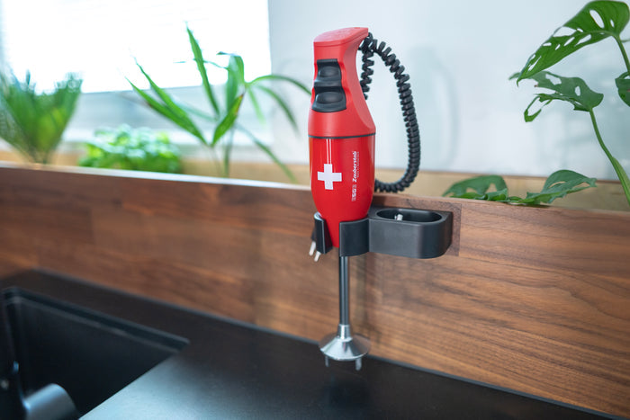 Der ESGE-Zauberstab E120 Select Premium-Set 902125 von ESGE Zauberstab, ein roter Stabmixer mit weißem Kreuz, ist an einer hölzernen Küchenwand in der Nähe von grünen Pflanzen und einer schwarzen Arbeitsplatte montiert.