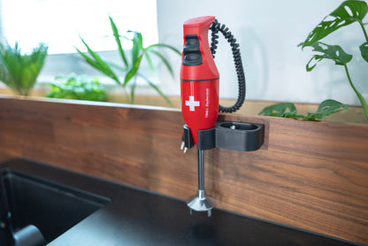 Der ESGE-Zauberstab E120 Select Premium-Set 902125 von ESGE Zauberstab, ein roter Stabmixer mit weißem Kreuz, ist an einer hölzernen Küchenwand in der Nähe von grünen Pflanzen und einer schwarzen Arbeitsplatte montiert.