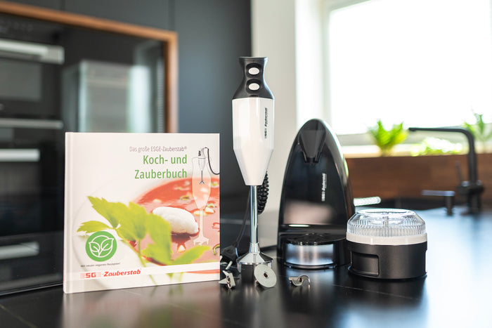 Der Stabmixer Unold ESGE-Zauberstab® Petticoat Edition mit Aufsätzen steht neben dem Koch- und Zauberbuch auf einer Küchentheke in einer sonnendurchfluteten modernen Küche.