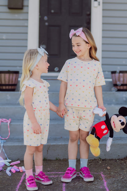 Two young girls in matching outfits standing on a set of stairs, holding hands with a Mickey Mouse plush toy.