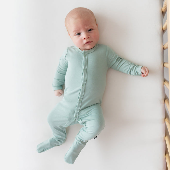 Newborn laying in crib and wearing Kyte Baby Footie in Sage