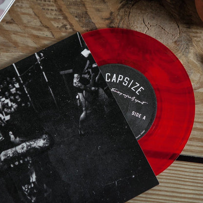 A limited edition transparent red vinyl from Capsize partially slides out of the black-and-white Ive Been Tearing Myself Apart album cover on a wooden surface; the label shows Capsize and Side A.