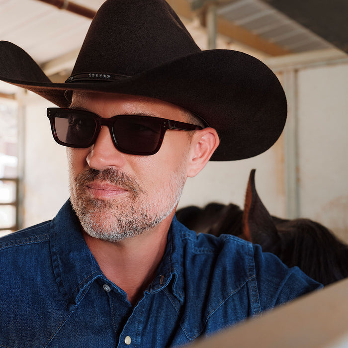 lifestyle - male in horse stables wearing yellowstone x diff eyewear john dutton rectangular sunglasses with a redwood frame and grey polarized lenses angled view