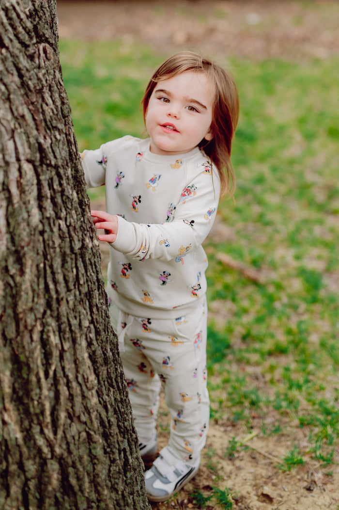 Child in a mickey mouse and friends patterned outfit standing next to a tree in an outdoor setting