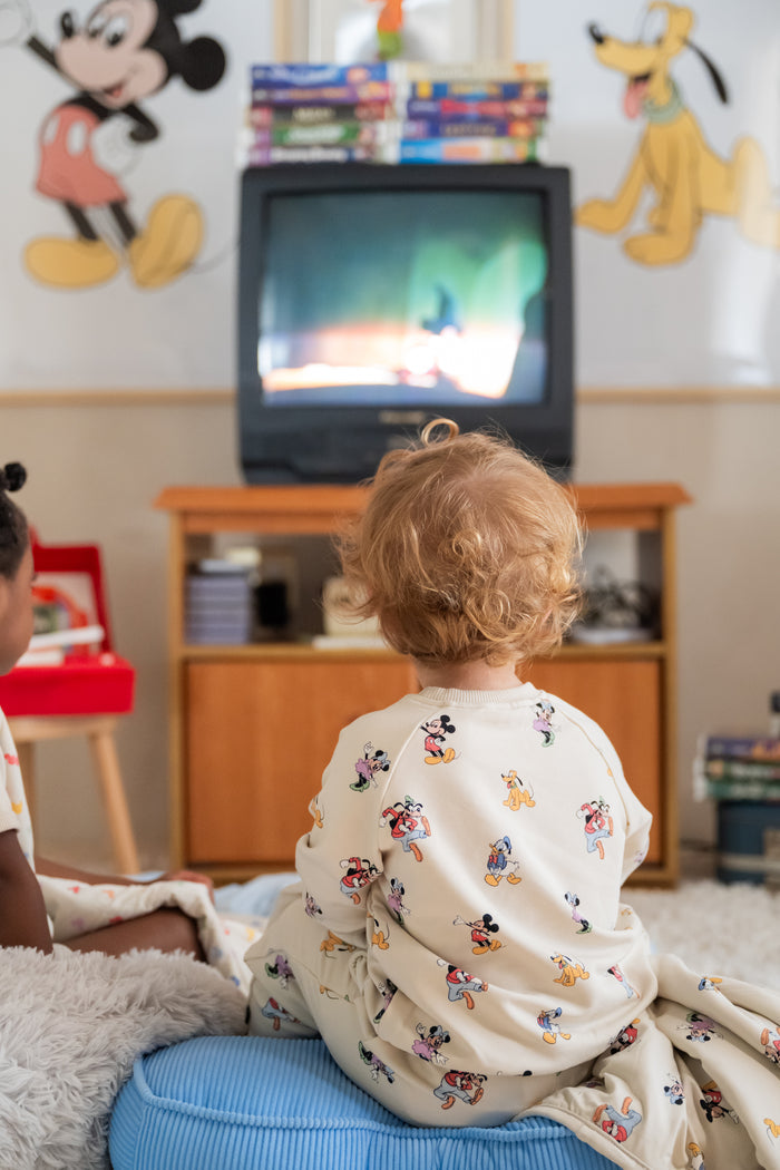 Two children sitting in a room with a television on a wooden stand, surrounded by toys and cartoon characters on the walls.