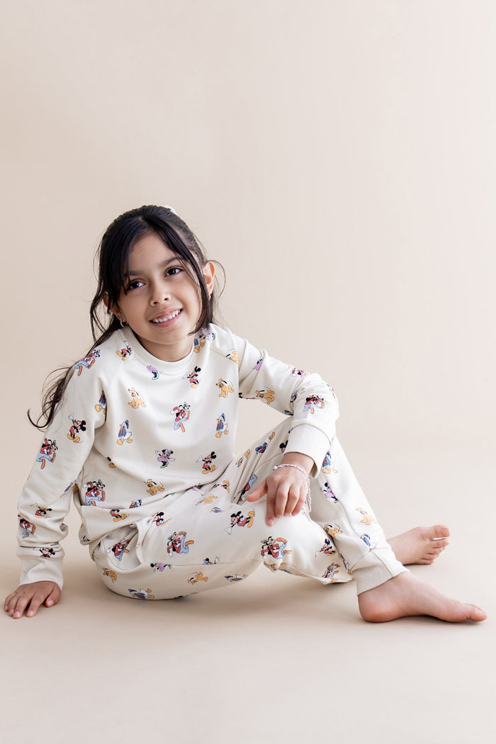 Child wearing a cream mickey character jogger set sitting on a beige surface.