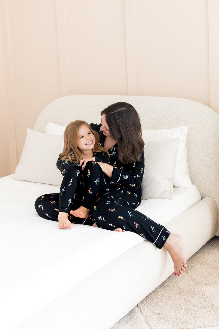 Model and girl wearing matching long sleeve pajama sets in midnight wildflower print sitting on white bed