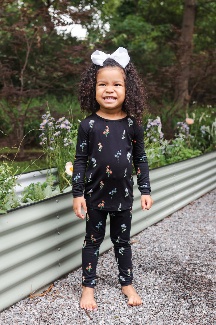 Girl wearing Long Sleeve Pajamas in Midnight Wildflower print and white head bow standing outdoors near plants