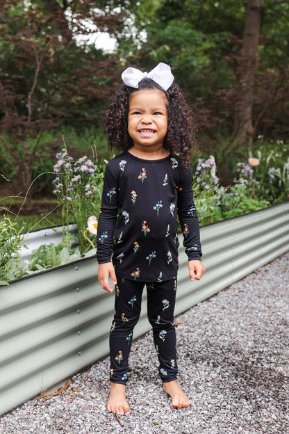 Girl wearing Long Sleeve Pajamas in Midnight Wildflower print and white head bow standing outdoors near plants