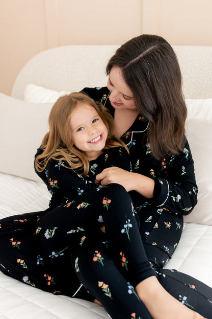 Model and girl sitting on white bed wearing matching long sleeve pajama sets in midnight wildflower print