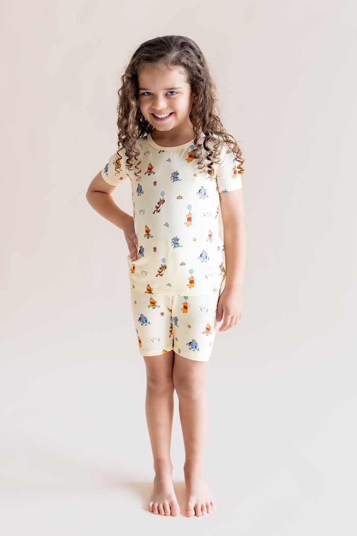 Girl wearing Short Sleeve Pajamas in Winnie the Pooh Celebration print posing on a cream background