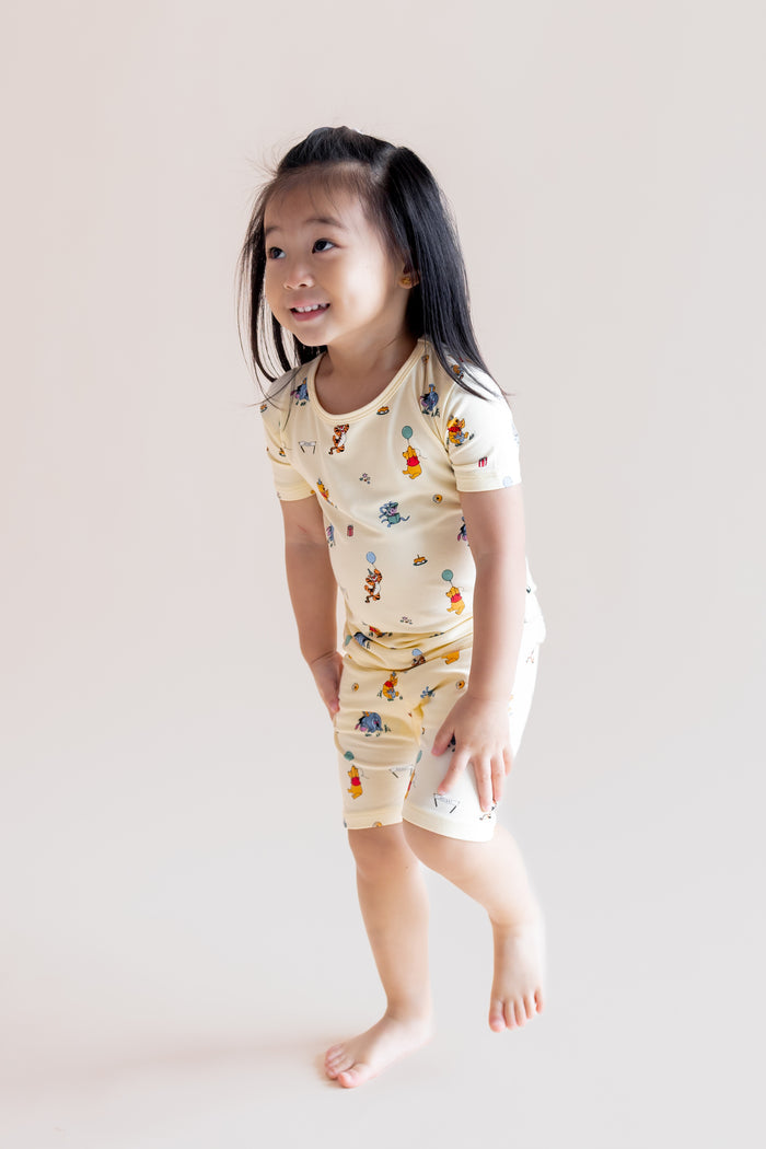 Girl standing wearing Short Sleeve Pajamas in Winnie the Pooh Celebration print on a cream background