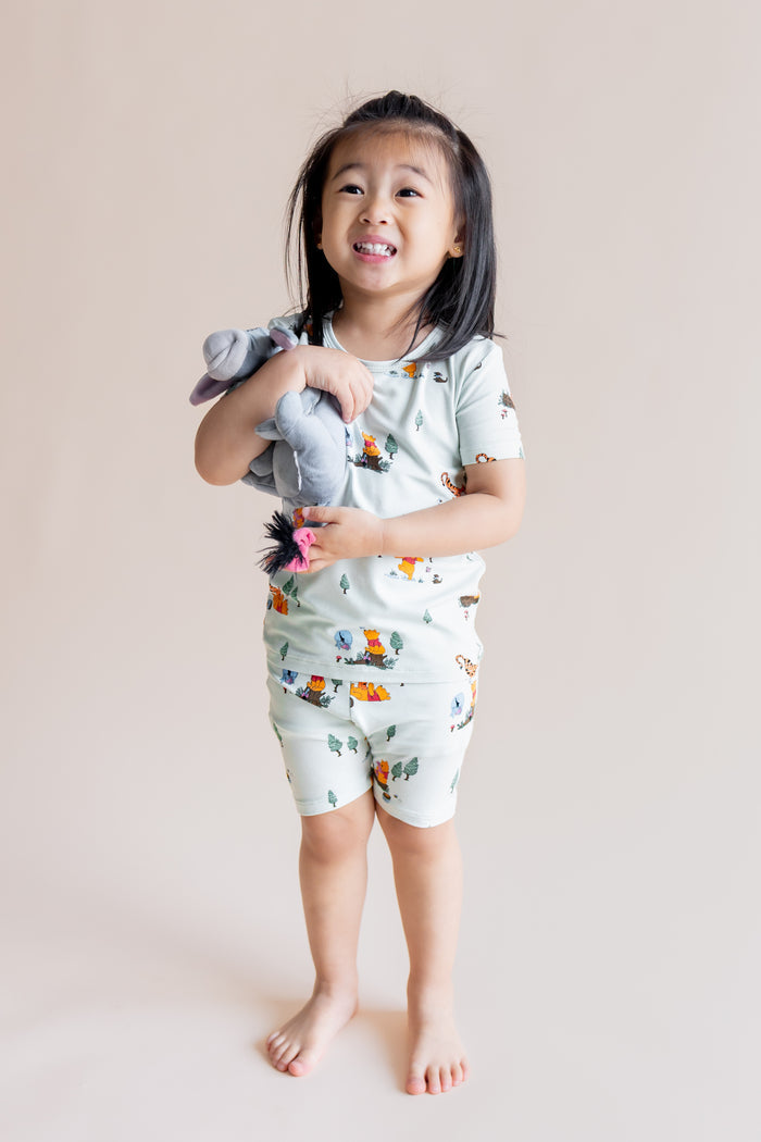 Girl smiling in Short Sleeve Pajamas in Winnie the Pooh Hundred Acre Wood print holding a stuff toy on a cream background