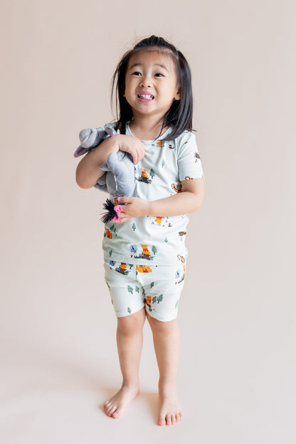 Girl smiling in Short Sleeve Pajamas in Winnie the Pooh Hundred Acre Wood print holding a stuff toy on a cream background