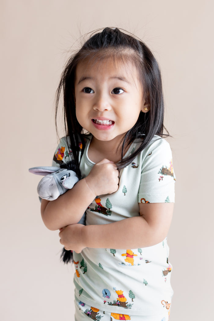 Girl holding a stuff toy wearing Short Sleeve Pajamas in Winnie the Pooh Hundred Acre Wood print on a cream background