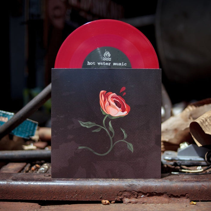 A red opaque vinyl record labeled Hot Water Music is partially pulled from a black sleeve with a red rose and green stem, displayed outdoors among scattered metal items—an ideal collector’s item for presale fans.