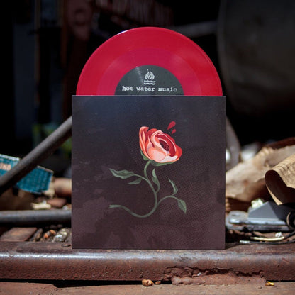 A red opaque vinyl record labeled Hot Water Music is partially pulled from a black sleeve with a red rose and green stem, displayed outdoors among scattered metal items—an ideal collector’s item for presale fans.