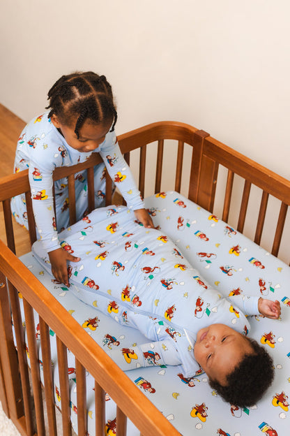 Baby lying in Crib in Sleep Bag in Curious George 1.0 print lying on a similar print crib sheet and a girl sitting outside the crib wearing Curious George print Sleep bag walker