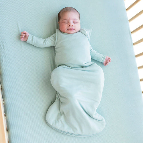 Infant sleeping in a crib and wearing a  Sleep Bag in Sage 2.5 with a long sleeve Sage romper underneath