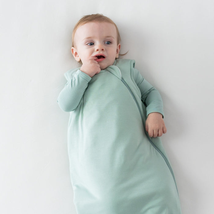Baby with finger in mouth wearing Kyte Baby Sleep Bag in Sage 2.5 and matching long sleeve bodysuit underneath