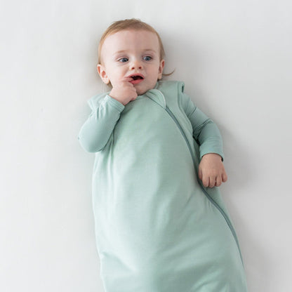Baby with finger in mouth wearing Kyte Baby Sleep Bag in Sage 2.5 and matching long sleeve bodysuit underneath