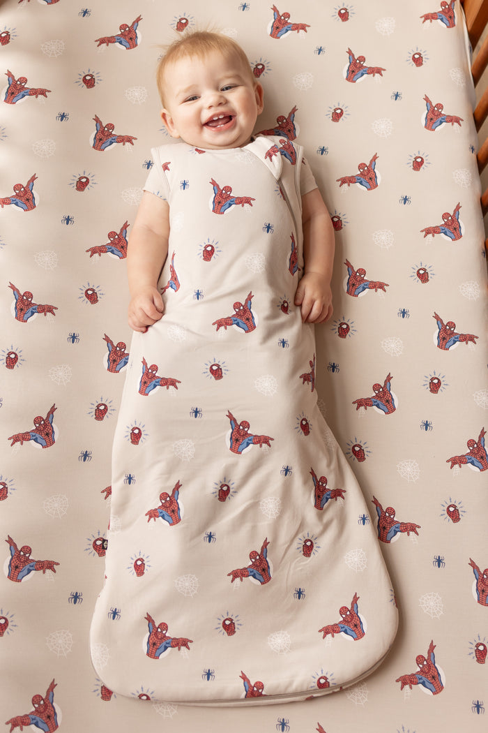 Baby in oat color Sleep Bag with Spider-Man print lying on a similar print crib sheet.