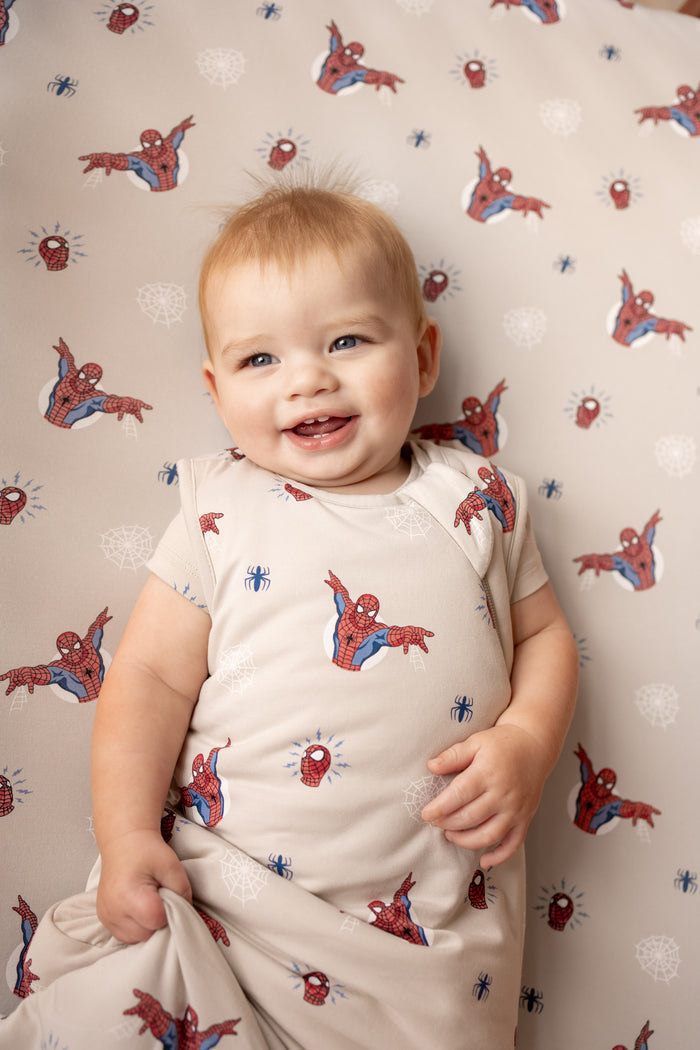 Baby smiling in oat color Sleep Bag with Spider-Man print lying on a similar print crib sheet.