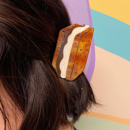 A s'mores shaped hair claw clip holding up brunette hair against a colorful background. The graham crackers have x's and o's on them in honor of the XO Marshmallow Brand.