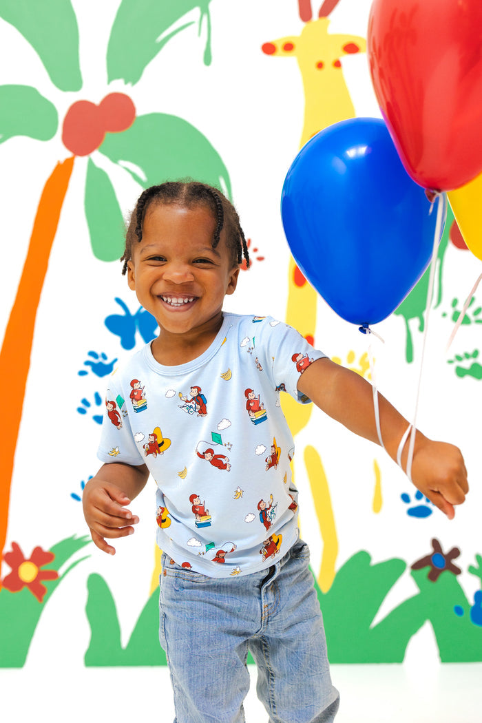 Girl wearing Toddler Basic Tee in Curious George print over a blue jeans holding red blue balloons in front of a colorful background