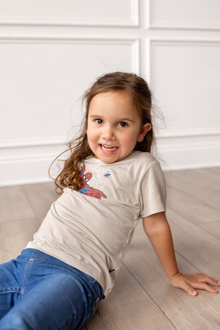 Girl on floor sitting in front of a wall wearing oat color Toddler Basic Tee with Spider-Man print in center over blue jeans