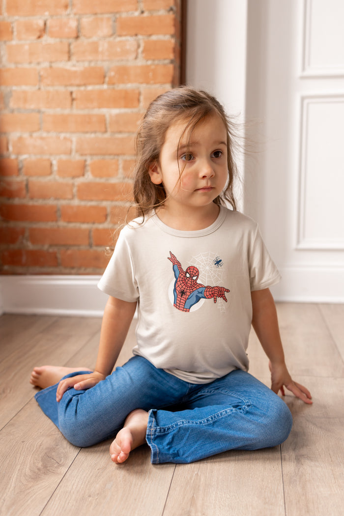 Girl posing on floor in front of a brick wall wearing oat color Toddler Basic Tee with Spider-Man print in center over blue jeans