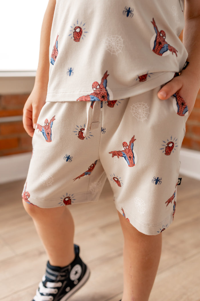 Boy wearing oat color Toddler Drawstring shorts under Toddler basic tee with Spider-Man print in front of a brick wall