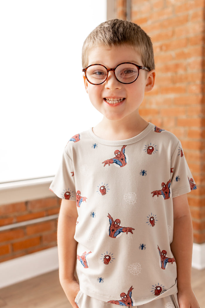 Boy in specs smiling in oat color Toddler Basic Tee with Spider-Man print in front of a brick wall