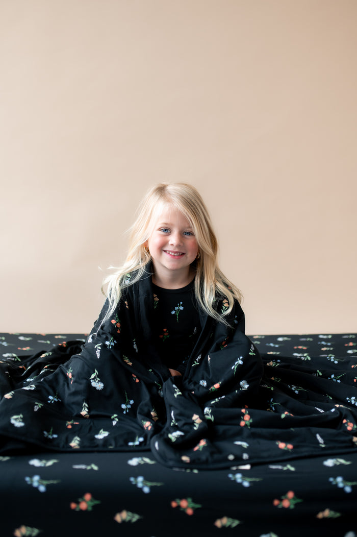 A girl wrapped in Toddler Blanket in Midnight Wildflower 1.0 print sitting on a similar print twin sheet and wearing similar print pajamas on a beige background