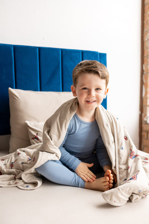 Boy sitting on bed wrapped in oat color Toddler Blanket with Spider-Man print wearing blue pajamas