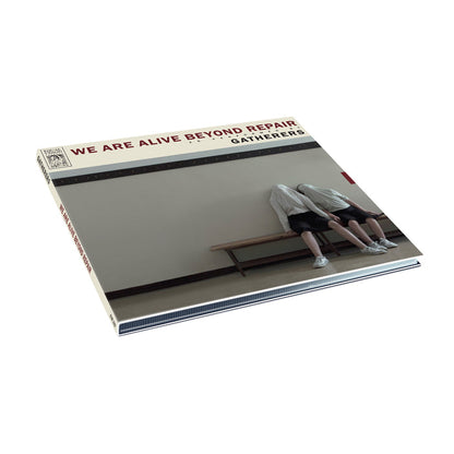 The We Are Alive Beyond Repair vinyl record cover by Gatherers features two people in Monochrome Coats, sitting on a bench with their heads covered by shirts, set against a plain wall.