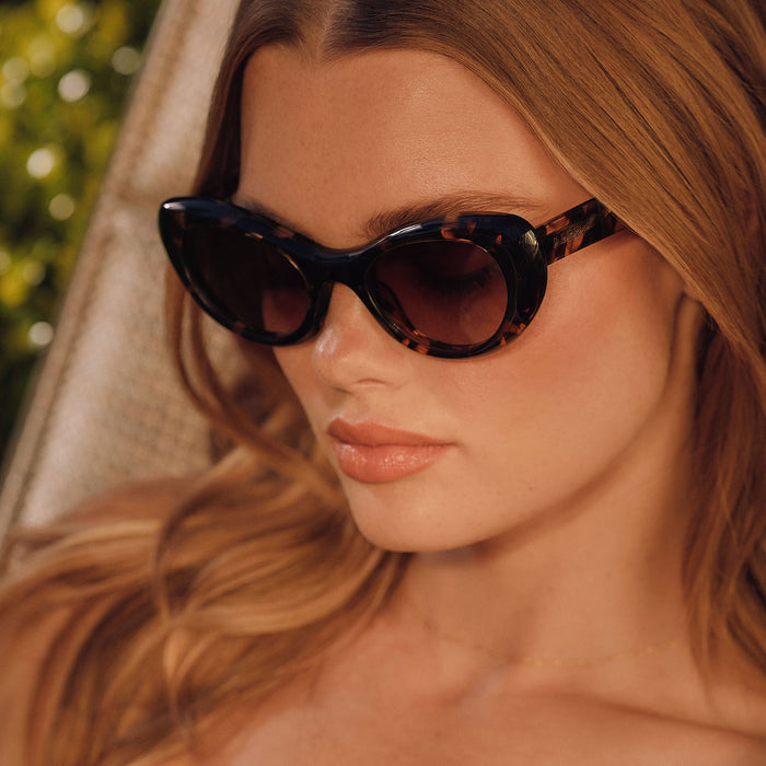 lifestyle - female lounging under some trees wearing diff eyewear zaria cat eye sunglasses with a espresso tortoise frame and brown gradient lenses angled view